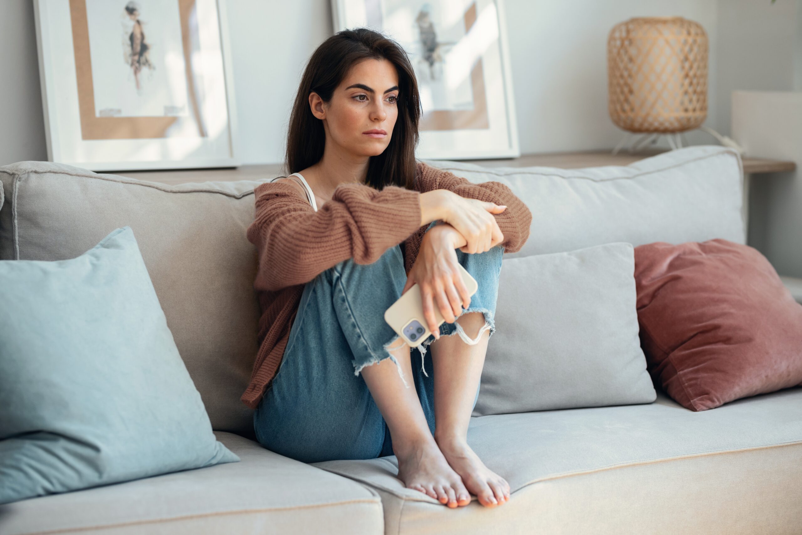 A woman sits thoughtfully on a sofa, holding her phone, reflecting the struggle of overthinking.